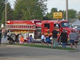 A large red fire truck with the words 'PIPE CREEK TWP FIRE RESCUE' is driving on a street. Several people, including children, are standing and sitting on folding chairs by the roadside, watching the fire truck. The scene is set in a suburban area with trees and houses in the background. A 'DOLLAR GENERAL' store sign is visible in the background.
