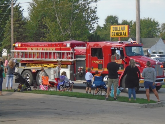 A large red fire truck with the words 'PIPE CREEK TWP FIRE RESCUE' is driving on a street. Several people, including children, are standing and sitting on folding chairs by the roadside, watching the fire truck. The scene is set in a suburban area with trees and houses in the background. A 'DOLLAR GENERAL' store sign is visible in the background.