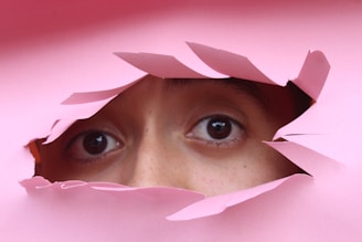 A pair of eyes peering through a torn hole in pink paper, creating a sense of curiosity and mystery.
