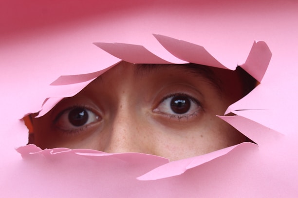 A pair of eyes peering through a torn hole in pink paper, creating a sense of curiosity and mystery.