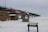 A bridge stretches across a snowy landscape, with a partially visible red sign indicating a Visitors Center and Fort Entrance. There's a small brown building with informational signs and a board displaying information for visitors. Snow covers the ground extensively, and the sky is overcast.