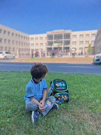 A young child sits on green grass near a colorful Minecraft-themed backpack. In the background, a large school building with many windows is visible, and several people can be seen entering or leaving the building. The sky is clear and blue.