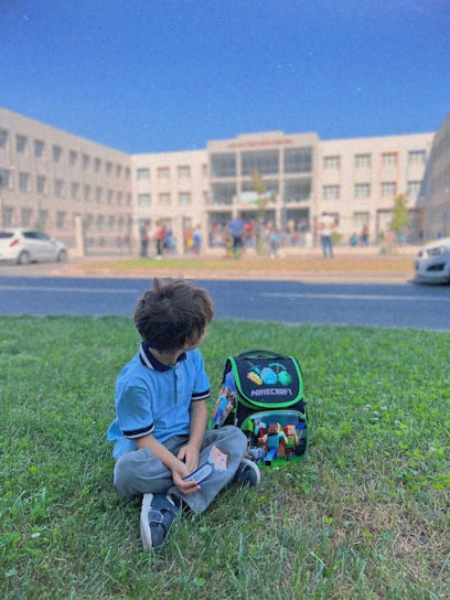 A young child sits on green grass near a colorful Minecraft-themed backpack. In the background, a large school building with many windows is visible, and several people can be seen entering or leaving the building. The sky is clear and blue.