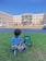 A young child sits on green grass near a colorful Minecraft-themed backpack. In the background, a large school building with many windows is visible, and several people can be seen entering or leaving the building. The sky is clear and blue.