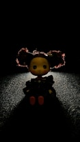 A small doll with large, curly hair sits on a textured surface. The doll is illuminated from above, creating dramatic shadows against the backdrop. Its hair is accented with red highlights, and it features minimal facial details.