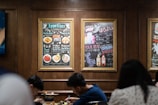 A cozy restaurant scene with two people sitting at a table, enjoying a meal. The walls have framed menus with colorful images and text in a combination of English and Japanese. The menus display various appetizers and drinks. The setting has warm wooden paneling and a casual dining atmosphere.