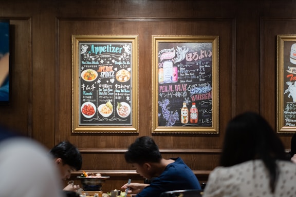 A cozy restaurant scene with two people sitting at a table, enjoying a meal. The walls have framed menus with colorful images and text in a combination of English and Japanese. The menus display various appetizers and drinks. The setting has warm wooden paneling and a casual dining atmosphere.