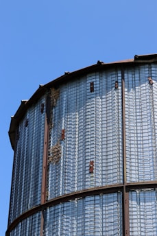 A close-up view of a metal grain silo with a grid structure and small tufts of grass or straw caught in the mesh. The silo is situated against a clear blue sky, and rust can be observed on the metal components.