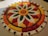 An intricate floral pattern made entirely of colored flower petals arranged on a flat surface. The design features concentric circular patterns with a central sun-like motif and alternating petal shapes radiating outward. The colors are vibrant and include shades of red, orange, yellow, green, and white, creating a visually striking and symmetrical display.