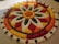 An intricate floral pattern made entirely of colored flower petals arranged on a flat surface. The design features concentric circular patterns with a central sun-like motif and alternating petal shapes radiating outward. The colors are vibrant and include shades of red, orange, yellow, green, and white, creating a visually striking and symmetrical display.