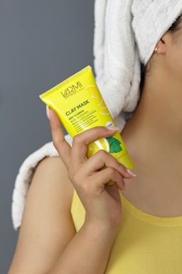 A person is holding a yellow tube of clay mask labeled 'VARMI' with a focus on mint and lemon. The person has a towel wrapped around their head and is wearing a yellow top, highlighting a skincare routine.