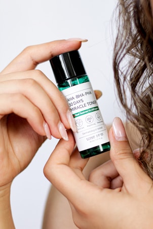 A hand holds a small bottle labeled with skincare information, while curly hair is visible in the background. The label mentions AHA, BHA, and PHA, indicating its use as a toner for skin care.