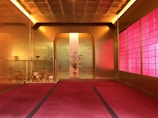 A room with a luxurious golden interior, adorned with opulent decor. On the left, a series of traditional Japanese tea utensils are displayed, including a teapot and cups. In the center, a vase with an arrangement of flowers adds elegance and natural beauty. The right wall features a series of translucent, reddish panels that diffuse the light.