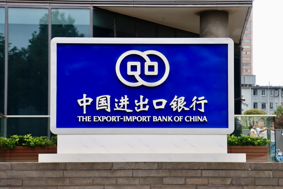 A large blue sign with white lettering and a logo displayed prominently. The text includes Chinese characters and English words, reading 'The Export-Import Bank of China'. The sign is situated in front of a modern building with glass windows and some greenery.