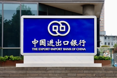 A large blue sign with white lettering and a logo displayed prominently. The text includes Chinese characters and English words, reading 'The Export-Import Bank of China'. The sign is situated in front of a modern building with glass windows and some greenery.