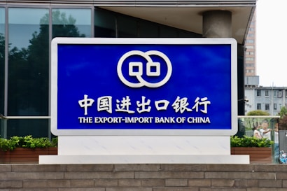 A large blue sign with white lettering and a logo displayed prominently. The text includes Chinese characters and English words, reading 'The Export-Import Bank of China'. The sign is situated in front of a modern building with glass windows and some greenery.