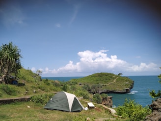 A serene coastal landscape with a small camping tent pitched on a grassy hill overlooking the ocean. The clear blue sky and white clouds enhance the peaceful atmosphere. Lush green vegetation and a rocky outcrop leading to a small cliff are visible in the distance.