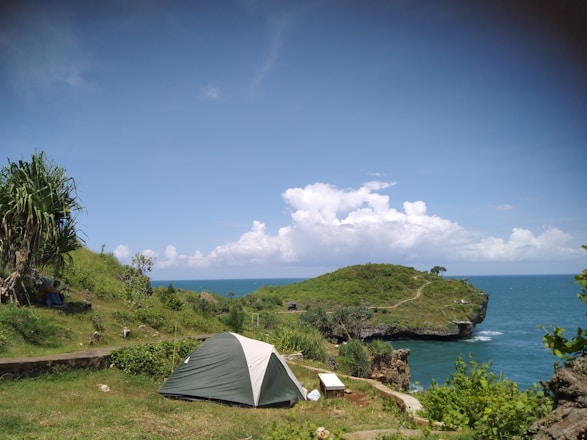 A serene coastal landscape with a small camping tent pitched on a grassy hill overlooking the ocean. The clear blue sky and white clouds enhance the peaceful atmosphere. Lush green vegetation and a rocky outcrop leading to a small cliff are visible in the distance.