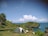 A serene coastal landscape with a small camping tent pitched on a grassy hill overlooking the ocean. The clear blue sky and white clouds enhance the peaceful atmosphere. Lush green vegetation and a rocky outcrop leading to a small cliff are visible in the distance.
