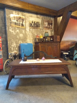 A rustic setting featuring a wooden table with decorative items on a lace table runner. Behind the table, there is a large wooden cabinet with vintage objects on top including jars and bottles. On the wall, there are three framed sepia-toned photographs. A vintage blue refrigerator stands to the left of the cabinet, and a red vending machine is partially visible. There is also a classic bicycle parked near the refrigerator.