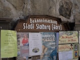 A public notice board displays several posters and announcements. These include a large wooden sign with 'Bekanntmachung Stadt Stolberg (Harz)' at the top. Below are various posters advertising local events such as a flea market, a book reading, and a cultural festival. The board is set against a stone wall.