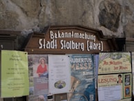 A public notice board displays several posters and announcements. These include a large wooden sign with 'Bekanntmachung Stadt Stolberg (Harz)' at the top. Below are various posters advertising local events such as a flea market, a book reading, and a cultural festival. The board is set against a stone wall.