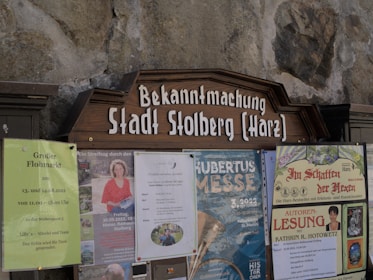 A public notice board displays several posters and announcements. These include a large wooden sign with 'Bekanntmachung Stadt Stolberg (Harz)' at the top. Below are various posters advertising local events such as a flea market, a book reading, and a cultural festival. The board is set against a stone wall.