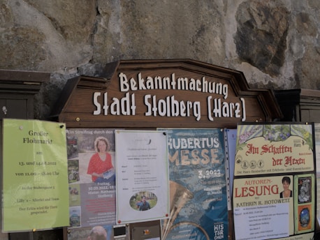 A public notice board displays several posters and announcements. These include a large wooden sign with 'Bekanntmachung Stadt Stolberg (Harz)' at the top. Below are various posters advertising local events such as a flea market, a book reading, and a cultural festival. The board is set against a stone wall.