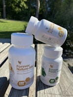 Three white plastic bottles of dietary supplements with labels. Two bottles are upright on a wooden surface, while the third is balanced on top. The background features green grass and trees in soft focus.