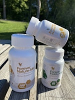 Three white plastic bottles of dietary supplements with labels. Two bottles are upright on a wooden surface, while the third is balanced on top. The background features green grass and trees in soft focus.