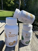 Three white plastic bottles of dietary supplements with labels. Two bottles are upright on a wooden surface, while the third is balanced on top. The background features green grass and trees in soft focus.