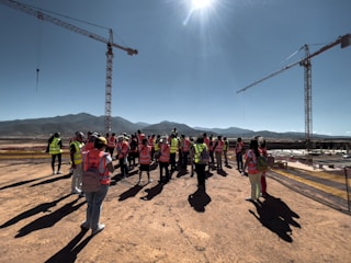 A group of people wearing high-visibility vests is gathered on a construction site. Two large cranes are visible in the background, along with distant mountains under a clear blue sky. The sun is bright overhead, casting long shadows on the ground.