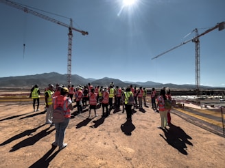 A group of people wearing high-visibility vests is gathered on a construction site. Two large cranes are visible in the background, along with distant mountains under a clear blue sky. The sun is bright overhead, casting long shadows on the ground.