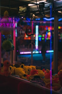 A claw machine filled with various colorful plush toys is seen inside an amusement arcade. Bright neon lights illuminate the background, casting vibrant reflections on the glass surfaces.