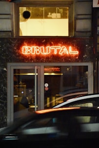 A storefront features a prominent red neon sign displaying the word 'BRUTAL.' Beneath the sign, there are glass doors and dark-colored marble walls. Reflections of lights and blurred movement suggest a street scene with passing cars.
