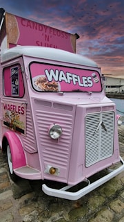 A vintage pink food truck with bright signage advertising waffles. The truck features an old-fashioned design with a prominent front grille and large, round headlights. The sign displays images of waffles topped with various toppings. The sky has a dramatic sunset with shades of pink and orange, and birds are visible flying in the distance.