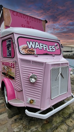 A vintage pink food truck with bright signage advertising waffles. The truck features an old-fashioned design with a prominent front grille and large, round headlights. The sign displays images of waffles topped with various toppings. The sky has a dramatic sunset with shades of pink and orange, and birds are visible flying in the distance.