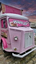 A vintage pink food truck with bright signage advertising waffles. The truck features an old-fashioned design with a prominent front grille and large, round headlights. The sign displays images of waffles topped with various toppings. The sky has a dramatic sunset with shades of pink and orange, and birds are visible flying in the distance.