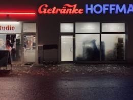 A storefront at night featuring two adjacent businesses. On the left, a hair studio with signs on the door and windows. On the right, a beverage store named 'Getränke Hoffmann' with illuminated red and blue signage. The sidewalk is covered with scattered leaves, and the reflections on wet pavement suggest recent rain.