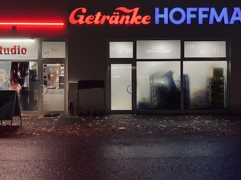 A storefront at night featuring two adjacent businesses. On the left, a hair studio with signs on the door and windows. On the right, a beverage store named 'Getr&auml;nke Hoffmann' with illuminated red and blue signage. The sidewalk is covered with scattered leaves, and the reflections on wet pavement suggest recent rain.