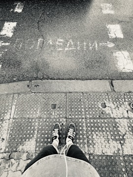 An overhead view shows a person standing on a textured sidewalk, wearing black sneakers with white laces and holding an object with a white cable, possibly earphones. The sidewalk leads to a cracked and worn road with partially visible white text in an unknown language and arrow markings.