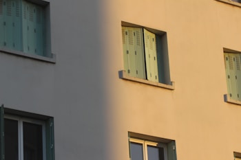 A building exterior with mint green window shutters that contrast with the beige walls. The light from the setting sun casts a shadow, splitting the facade into light and dark sections.