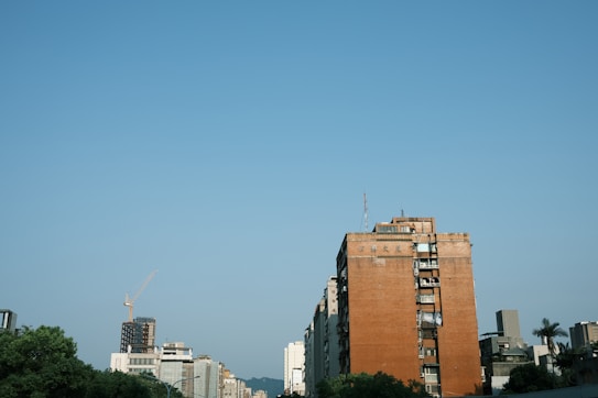 A cityscape featuring a prominent brick building with several windows and antennas on the roof. Other modern buildings, including a partially constructed tower with a crane, are visible in the background. The sky is clear and blue, with a cluster of trees in the foreground adding greenery.