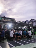 A group of people seated at tables under a covered outdoor area, presumably engaged in a communal meal or gathering. The setting appears to be in a parking lot or courtyard, with modern buildings in the background and a somewhat cloudy sky overhead.