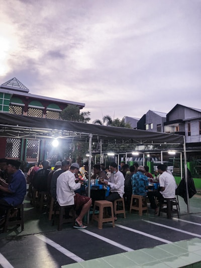 A group of people seated at tables under a covered outdoor area, presumably engaged in a communal meal or gathering. The setting appears to be in a parking lot or courtyard, with modern buildings in the background and a somewhat cloudy sky overhead.