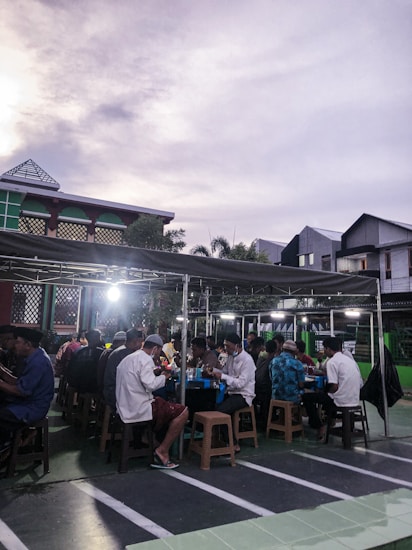 A group of people seated at tables under a covered outdoor area, presumably engaged in a communal meal or gathering. The setting appears to be in a parking lot or courtyard, with modern buildings in the background and a somewhat cloudy sky overhead.