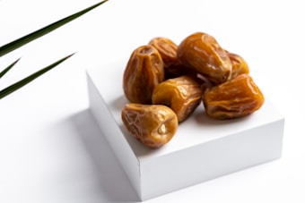 A cluster of brown dates is placed on a simple, white, cubical platform. The focus is on the glossy texture of the dates, with a few sharp leaves visible on the left side, suggesting a natural or organic theme.