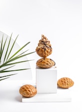 A stack of pastries topped with a swirl of cream and chocolate sauce, elegantly arranged on a white cube display. Surrounding the stack are several similar pastries placed on a clean white surface. A decorative green palm leaf is positioned to the left, adding a touch of natural color and contrast to the minimalist background.