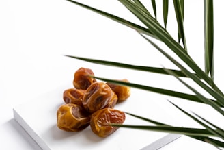 A cluster of glossy brown dates sits on a white rectangular surface, with some green palm fronds partially obscuring the view from the side.
