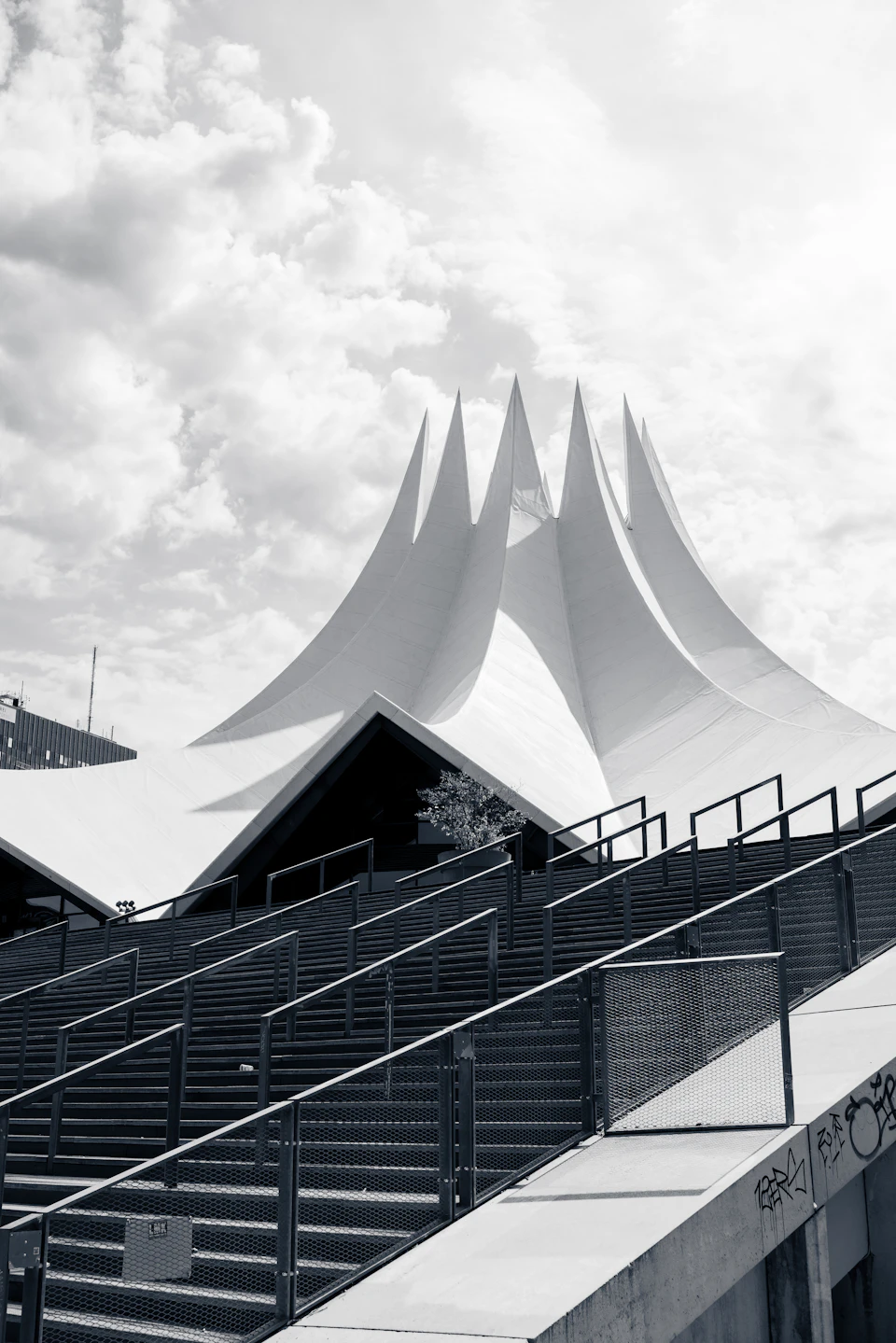 The image depicts a large, white building with a pointed roof and multiple spires, standing tall against an overcast sky. The building is situated on a concrete platform surrounded by stairs leading up to it. A railing can be seen in front of the building, providing safety for pedestrians. In the foreground, there are several steps that lead up to the entrance of the building.